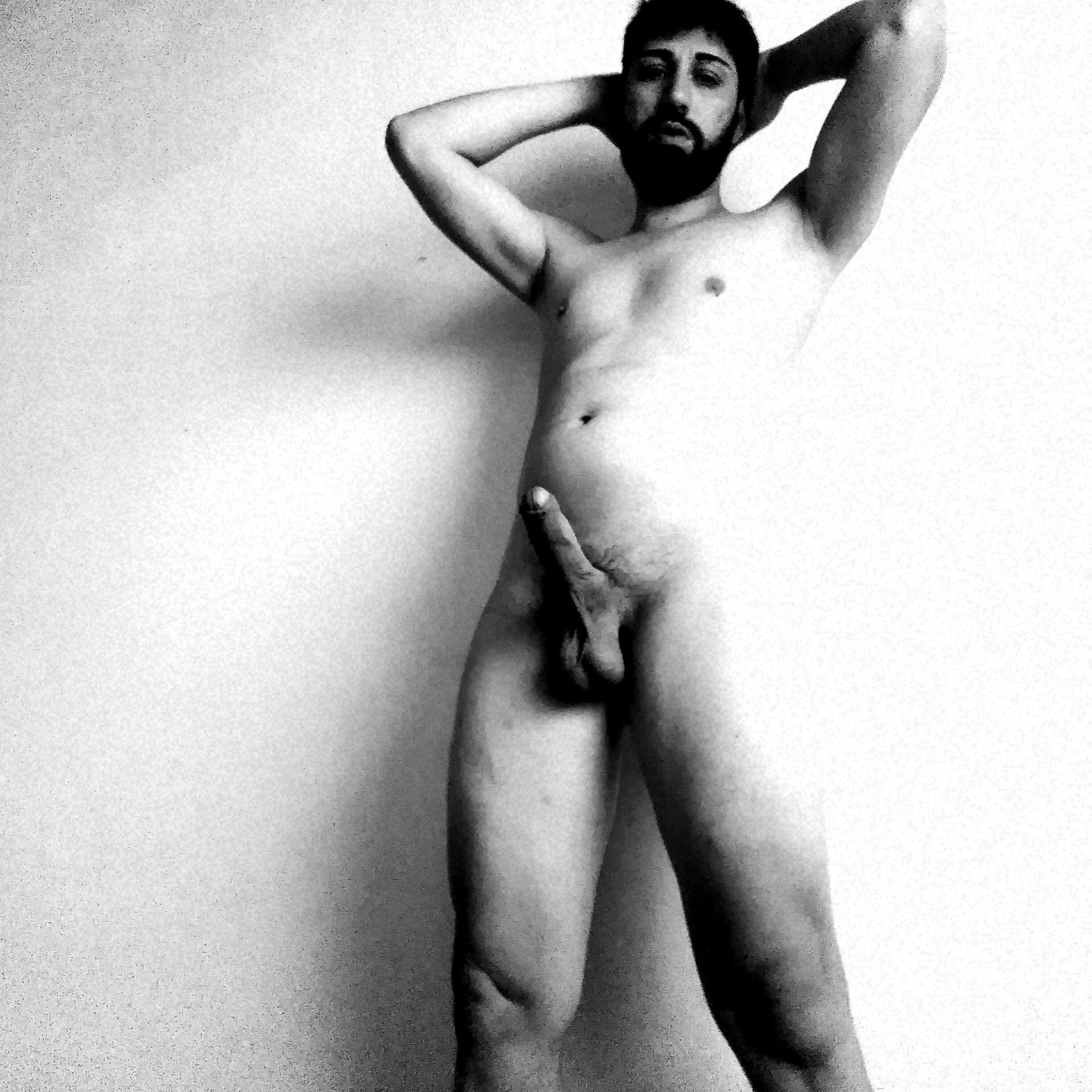 Hombre barbudo desnudo posando frontalmente con manos tras la cabeza y el pene erecto.